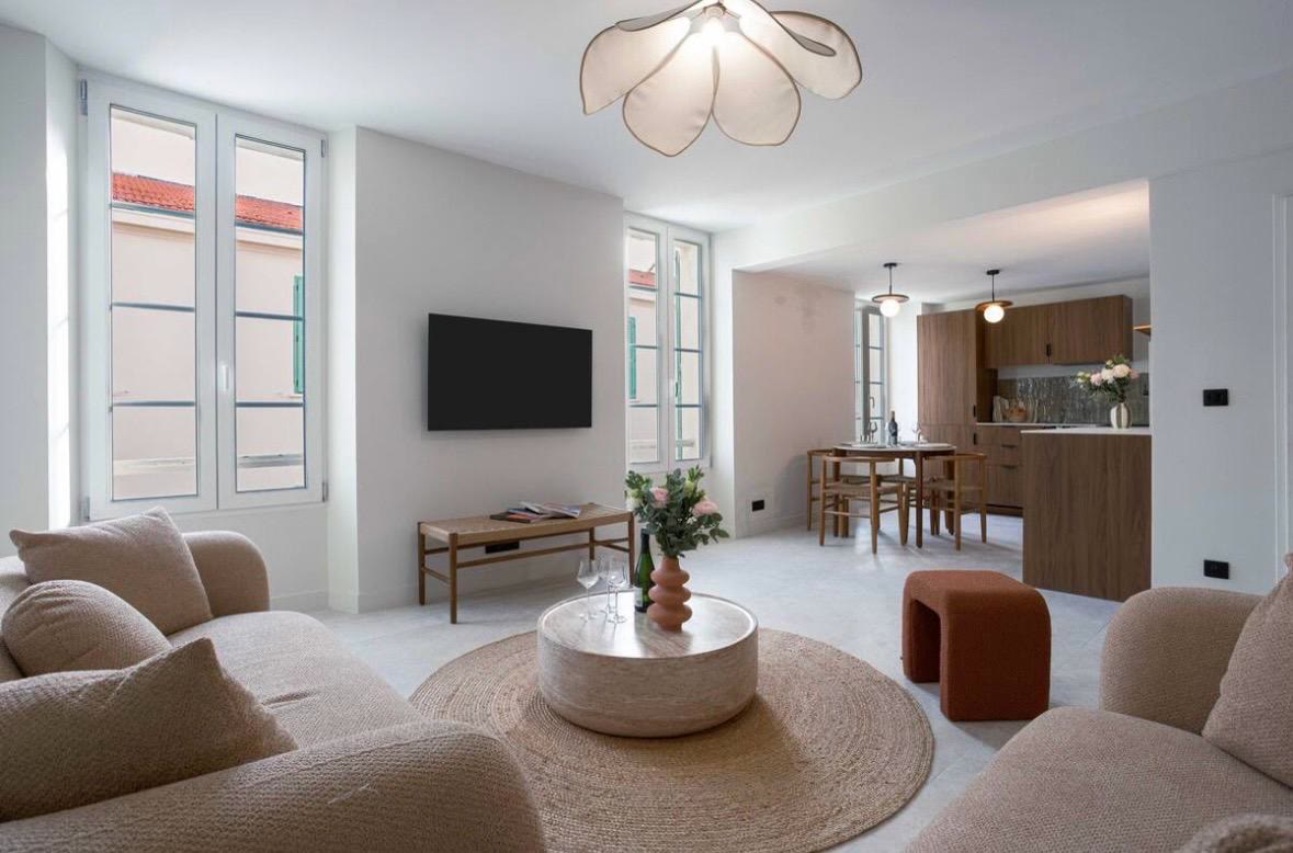 Le salon lumineux de L'Unique Jean Daumas, ses canapés en bouclette beige et sa cuisine ouverte en bois sous une grande suspension pétale.